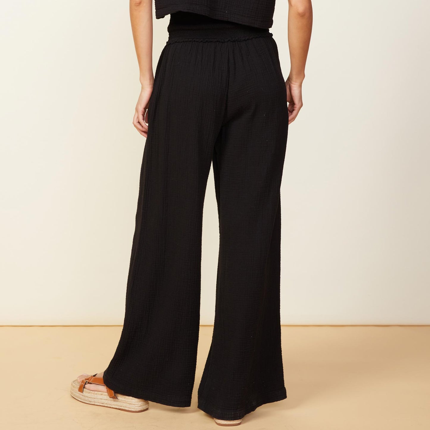 Back view of model wearing the gauze smocked flare pant in black.