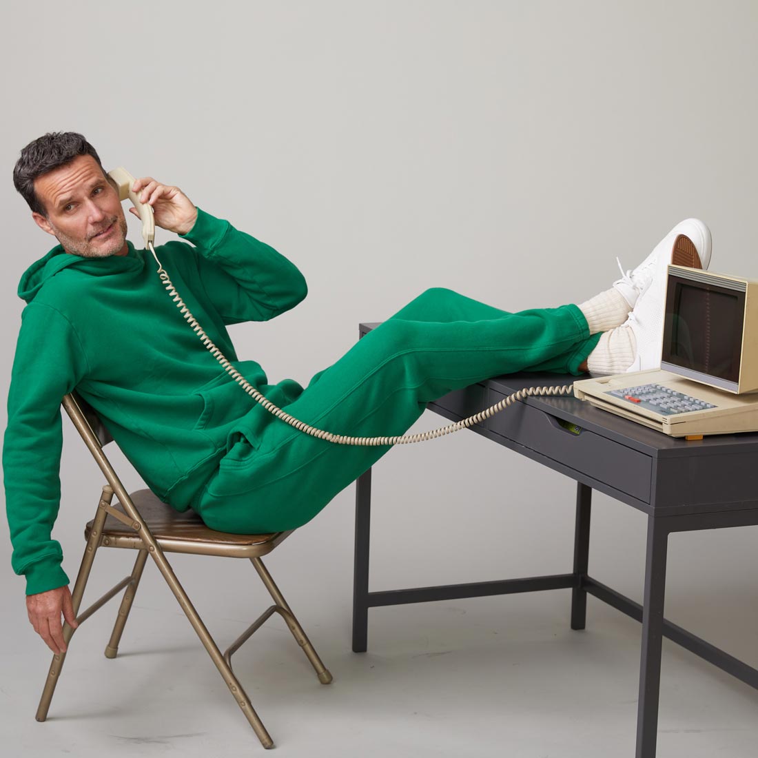 Model is sitting in a chair with his feet up on the a desk wearing the 70's sweats in dusty kelly while talking on a phone.